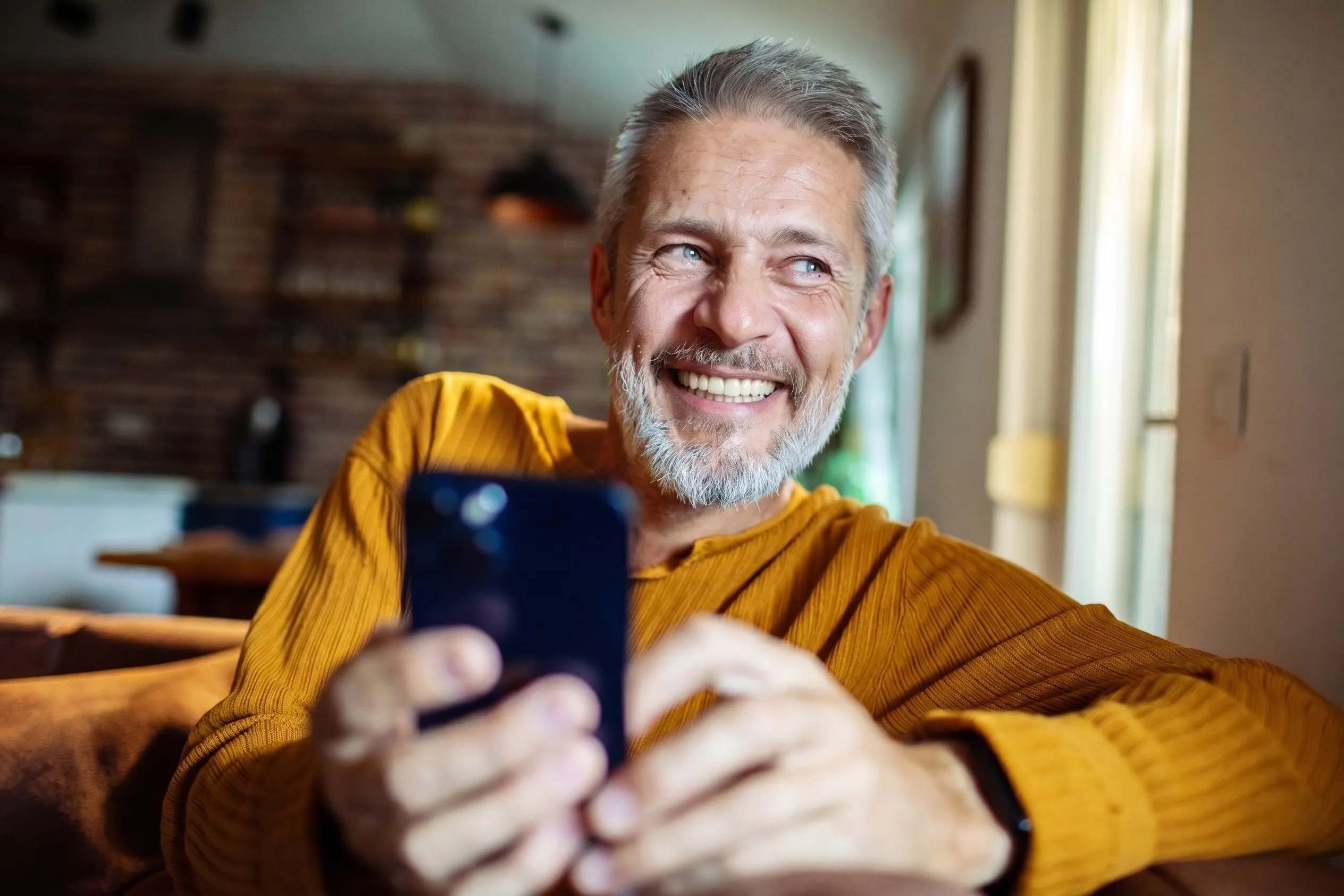 A relaxed older man smiling at home while holding his phone, in soft natural light
